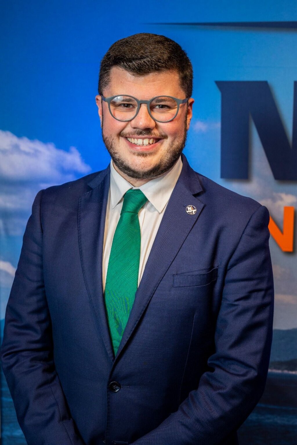 A picture of a smiling man, here is wearing a navy suit and green tie. He has dark hair and wears glasses.